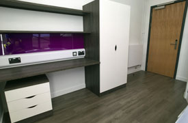 A room with a desk with nearby powerpoints and drawers underneath. There are two shelves over the desk and a cupboard next to it.
