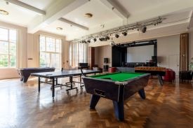 A large room with two pool tables, a ping pong table, and a table football table. At the back of the room there is a raised stage with a screen and a drum kit.