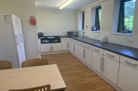 A room with a table with three chairs around it, two fridges, a fire blanket, a sink and lots of cupboards and drawers.