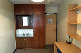 A room with a desk, chair and set of shelves against one wall. Against another wall there is a set of cupboards surrounding a sink with a mirror over it.