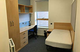 A room with a single bed in one corner and a desk against the opposite wall. The desk has an office chair nearby, a set of shelves above it, and a wardrobe to its side.