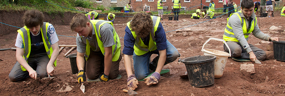 Groups of students carefully digging fenced-off rocky ground on an urban archeology site.
