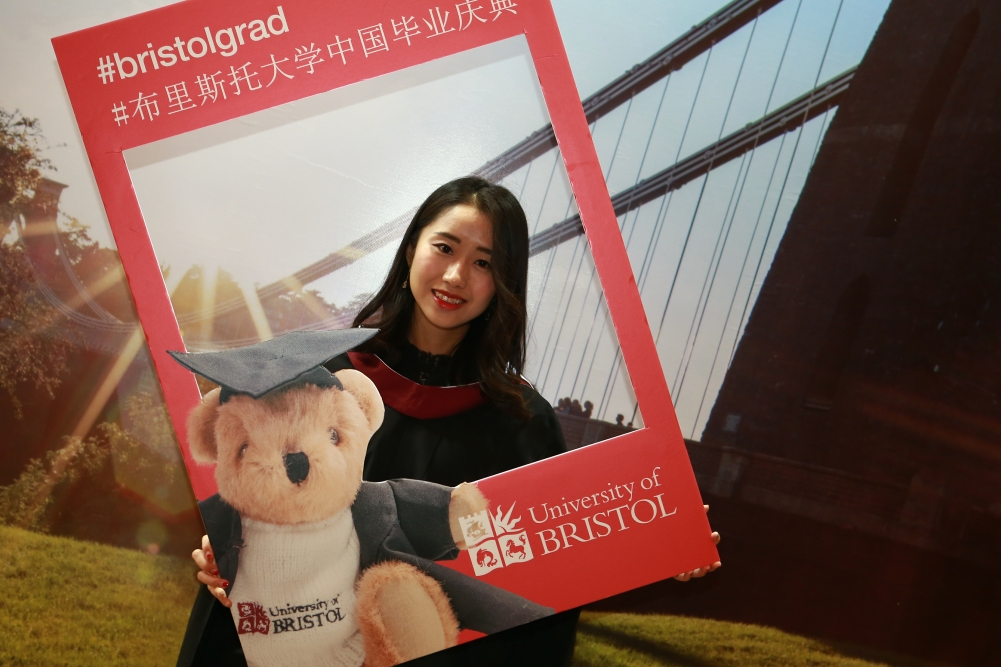 A Chinese graduate holds the #bristolgrad selfie photo frame in front of the Clifton Suspension Bridge.