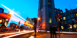 Lights from passing cars make solid lines appear against a backdrop of old University buildings and the dark blue night sky. 