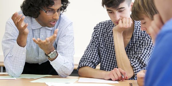 A man explains something to three people, who are sitting around a table working together. 