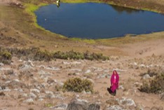 A young girl in flowing pink robes walks up the slope in front of a small mountain lake in Tanzania