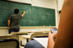 A student in the foreground is messaging on a mobile phone, whilst a teacher in the background writes on a blackboard