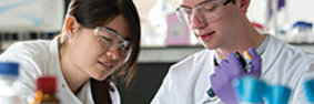 A woman and a man of different races in safety glasses and lab coats working together in a laboratory.
