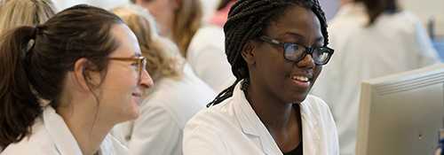 Two students in lab coats looking at a pc monitor screen. 