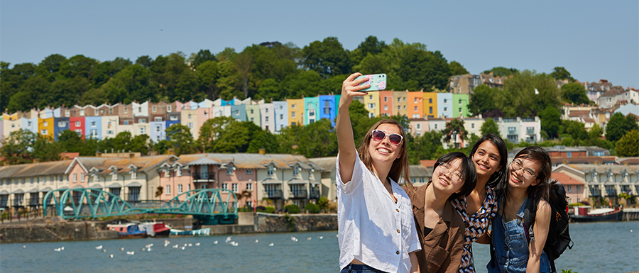A group of 4 international students taking a selfie together on a sunny day at Bristol harbour with colourful houses in the background. 