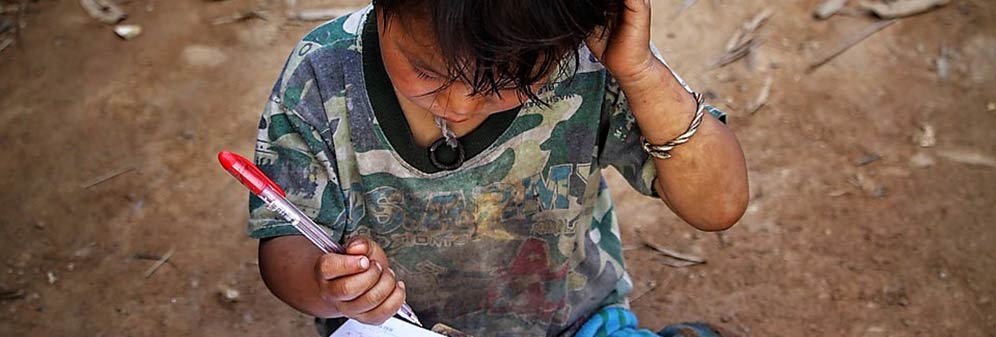 A child wearing dirty clothes and holding a pen, looking down at a piece of paper.