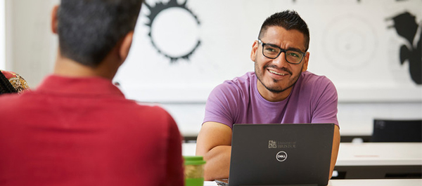 2 students sat at a table having a conversation with their laptops open.