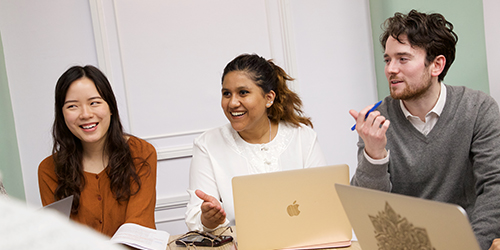 Three students with their laptops open smile while engaging in a conversation.