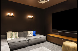 A cosy cinema or media room with dark walls, a large sectional sofa with patterned cushions, soft wall lighting, and a large TV screen on a low cabinet.