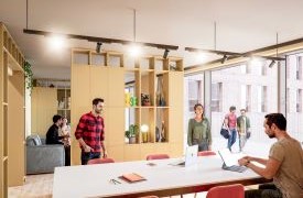 A student is sitting at a table in a shared communal space. There is a bookshelf and more students walking by.