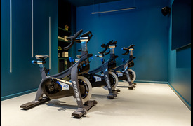 An exercise room with several stationary spin bikes arranged in a row against deep blue walls, lit by overhead lighting. A large screen, faced by the bikes, is on one wall.