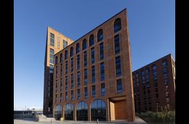 A modern multi‑storey red brick building with large rectangular and arched windows, under a bright blue sky. A tall vertical sign on the left side reads &ldquo;UNITE STUDENTS.