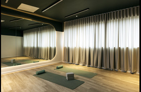 A calm fitness studio with wooden floors, soft lighting, and floor‑to‑ceiling curtains. Two yoga mats with rollers and blocks are laid out beside a large wall mirror.