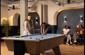 A games room featuring a pool table where two beaming people are playing, while two others sit on a bench against the wall, in comfortable conversation. The space has warm lighting, arched wall recesses, plants, and exposed ceiling fixtures.