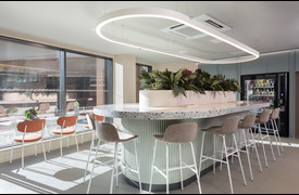 A bright caf&eacute;-style space with a curved terrazzo counter topped with plants, surrounded by high stools. Large windows line the room, and a vending machine is visible on the right.