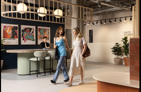 A modern reception area with two people, one joyful, the other somber, walking past a curved counter where another person is seated, frowning at a laptop. The space features abstract artwork on the wall, round hanging lights, indoor plants, and a sign reading &ldquo;Unite Students.&rdquo;