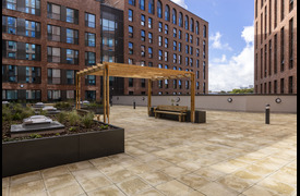 A spacious rooftop terrace surrounded by modern brick buildings, featuring paved flooring, planted garden beds, and a wooden pergola with bench seating under a bright sky.