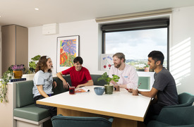 A group of four individuals sit around a white table in a modern shared kitchen space, in amicable conversation. The room includes built‑in green seating, several potted plants, and framed artwork on the wall. A large window behind the group shows an outdoor city view.