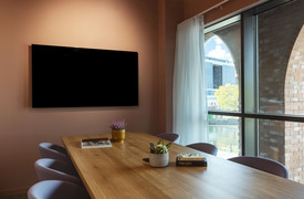 A room with a long with a long wooden table surrounded by modern chairs. A large black screen is mounted on the pink wall at the table's end. To the right, tall windows with sheer curtains let in natural light. A large window behind the group shows an outdoor city view.