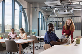 A modern study space with large arched windows, showing several people sitting at tables with books and laptops. They look like they are both studying and also enjoying themselves. The room features industrial-style lighting, soft-colored chairs, plants, and natural light streaming in from the windows.