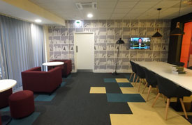 A room with a long table surrounded by chairs, and two smaller tables surrounded by armchairs and stools. There is a television of the wall at the end of the long table.
