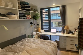 Bed and shelving units in the left foreground. Window and desk area in the background.