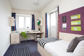 A room with a single bed in one corner and a desk and chair opposite it. There are two sets of shelves over the desk and drawers beneath it.