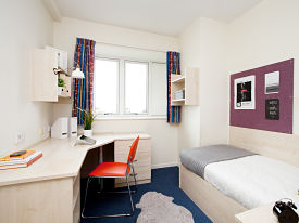 A room with a single bed in one corner and a desk and chair opposite it. There are two sets of shelves over the desk and drawers beneath it.