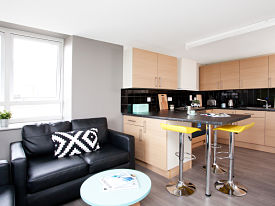 A room with a black leather sofa and a coffee table. The kitchenette area has a sink, and oven and hop, lots of cupboards, and two chairs at the kitchen counter.