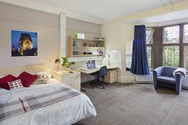 A room with a double bed in one corner and a desk next to it. The desk has an office chair beside it, drawers beneath it and shelves above it. There is also an armchair by the room's window.
