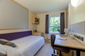 A room with a single bed against one wall and a desk and office chair opposite. There is a small chest of drawers at the foot of the bed and a set of shelves on the wall above it.