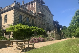 Exterior of a large four storey sandstone building. There is a garden in front of it with a bench and some steps leading up to the first floor of the building.