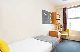 A room with a single bed in one corner and a desk with a chair opposite. There is a set of shelves on the wall above the desk and some drawers beneath it.