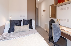 A room with a double bed in one corner and a desk with an office chair opposite. There is a cupboard behind the bed and a mirror on the wall opposite.
