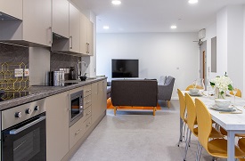 A kitchen with an oven, microwave, sink and lots of cupboards, with a dining table nearby with eight chairs around it. At the back of the room there is a television on the wall and several sofas near it.