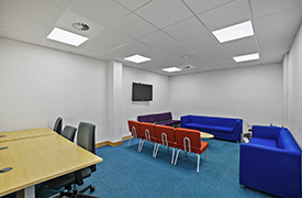 A room with a desk and three office chairs by it, a television on the wall, and two sofas and eight armchairs arranged around a coffee table nearby.