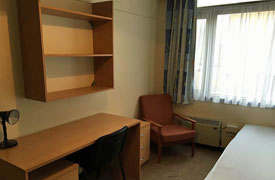 A room with a bed in one corner, and a desk and armchair against the opposite wall. The desk has another chair next to it, a set of shelves over it and a set of drawers beneath it.
