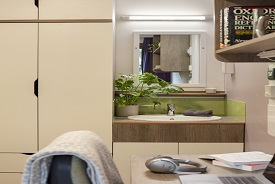 A room with a desk and office chair, a set of shelves over the desk, and a sink with a mirror over it and a wardrobe next to it.