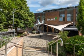 Steps leading down to a paved courtyard with trees, bushes and benches. At the end of the courtyard is a modern red brick two-storey building with a spiral staircase leading up to the first floor from the courtyard, and the word 'source' written on the wall.