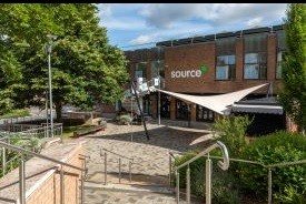 Steps leading down to a paved courtyard with trees, bushes and benches. At the end of the courtyard is a modern red brick two-storey building with a spiral staircase leading up to the first floor from the courtyard, and the word 'source' written on the wall.