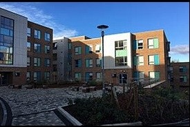 A paved courtyard with bushes and benches, enclosed on two sides by a four-storey modern red brick building.
