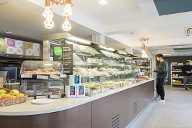 A man stood at the counter of a food shop. In front of him there are lots of sandwiches, some cake and some fruit.