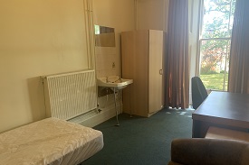 A bedroom with a desk, radiator, sink and a large window at the end of the room.
