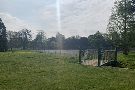 A lawn with a bridge going over a small gap, leading towards a set of tennis courts surrounded by a tall fence.
