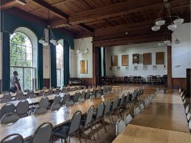 Large dining hall with long wooden tables and chairs. There are tall arched windows, and a raised stage area at the far end of the room.